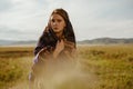 Mysterious girl in an ethnic cape anxiously looks at the camera, sitting in the grass in a field, in the steppes against