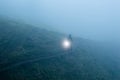 A mysterious hooded figure, carrying a glowing light. On a mountain path on a spooky foggy, winters evening.