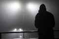 A mysterious hooded figure, standing with back to camera on a bridge, looking out at street lights. On a foggy night Royalty Free Stock Photo