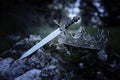 mysterious and magical photo of silver king crown and sword over the stone in the England woods or field landscape. Medieval