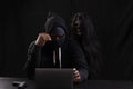 Mysterious man in hood with skull inside using laptop on table with gun on isolated black background. Hacker male hoodie