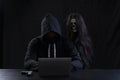 Mysterious man in hood with skull inside using laptop on table with gun on isolated black background. Hacker male hoodie