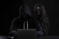 Mysterious man in hood with skull inside using laptop on table with gun on isolated black background. Hacker male hoodie