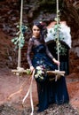 Mysterious woman in a black dress on a swing with flowers. Ruins of an old monastery. Black magic, Gothic beauty, mystical image,