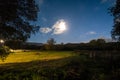 Mystical night field with a bright moon, moving clouds, and a light trail in the starry sky, with warm light on the grass Royalty Free Stock Photo
