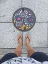 Top view of female legs wearing slippers in front of a colored manhole in Matsumoto, Japan