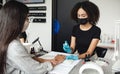 Nail salon during quarantine. African american girl in protective mask disinfects hands of client