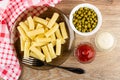 Napkin, bowls with green peas, mayonnaise, ketchup, brown plate with cooked pasta, fork on table. Top view