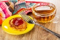 Napkin, cup with tea, cheese pancake with jam in saucer, fork on wooden table