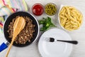 Napkin, spoon in frying pan with fried mince, bowls with green peas, ketchup, bowl with pasta, parsley, fork in plate on table.