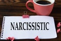 NARCISSISTIC - word on a white sheet on a wooden brown background with a cup of coffee and a pen