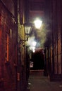 Narrow city alley illuminated by old glowing street lights shining on brick walls with atmospheric swirling fog and dark shadows Royalty Free Stock Photo