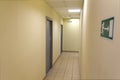 A narrow light corridor in the office room with several gray doors and signs.