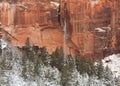 A narrow waterfall streams down a red sandstone cliff face in front of snow covered hills and pine trees Royalty Free Stock Photo