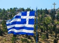 The national flag of Greece on the flagpole develops in the wind against the background of a hill with green trees and a cross on
