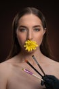 Natural cosmetics concept. Beauty model with bare shoulders holding a flower in her mouth and brushes in front looking Royalty Free Stock Photo