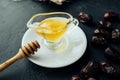 Natural honey in transparent saucepan with spout and wooden honey stick on saucer on dark table with dried dates scattered around