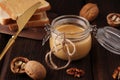 Natural nut butter in a glass jar, a golden knife, walnuts and sandwiches with toast on a wooden brown background. Vegetarian