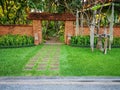 Natural orange clay brick arched wall entrance in a tropical garden with pattern of brown laterite walkway on green grass lawn