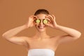 Natural skin detox. Charming young woman holding cucumber slices in front of her eyes on brown studio background Royalty Free Stock Photo