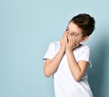 Naughty joyful boy in white t-shirt and jeans looking aside in surprise, covering his mouth with both hands. Royalty Free Stock Photo