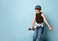 Naughty schoolboy in stylish cap jumping in place looking down under his feet with roguish smile. isolated on blue background