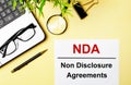 NDA Non Disclosure Agreements is written in red on a white piece of paper on a light yellow background next to a laptop, pen, Royalty Free Stock Photo