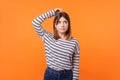 Need to think. Portrait of confused young woman with brown hair in long sleeve striped shirt. isolated on orange background