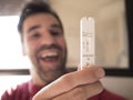 A negative covid test is held in his hand by a man with black hair and a short beard, out of focus, happy with his mouth open,