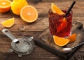 Negroni cocktail in crystal glass with orange slice and fresh raw oranges on chopping board with strainer on wooden background