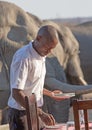 A waiter setting the table for guests in a safari lodge with a herd of elephants close by - Nehimba, Hwange National Park,