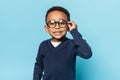 Nerdy african american boy in school uniform touching eyeglasses, looking and smiling at camera on blue background