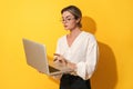 Nerdy woman wearing eyeglasses is using laptop computer on yellow background