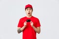 Nervous asian delivery guy in red t-shirt and cap uniform, holding credit card, staring camera and gasping. Young