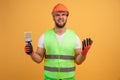 Nervous construction worker holds paint brush, looks aside, wears safety hardhat, dressed in reflector waistcoat, puzzled with