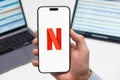 Netflix logo on the screen of a cell phone in mans hand and laptop on the background