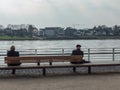 Neuwied, Germany - March 23, 2021: two men seen from behind, sitting with distance on a bench at the quayside of the Rhine