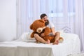 Never grow up concept. Guy on happy face hugs giant teddy bear. Man sits on bed and hugs big toy, white curtains on Royalty Free Stock Photo