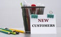 NEW CUSTOMERS - word on a white sheet with clips against the background of cans of pencils