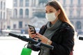 New eco-friendly transport. Young business woman with surgical mask unlocks e-scooter with her mobile phone. Woman uses an app on