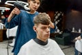 New haircut is almost done. Young stylish guy sitting in armchair in the modern barbershop or salon while professional Royalty Free Stock Photo