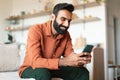 Cheerful Indian bearded man messaging on cell phone sitting indoor Royalty Free Stock Photo