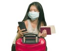 New normal traveling - young happy and beautiful Asian Korean woman in face mask carrying suitcase holding passport using mobile Royalty Free Stock Photo