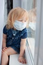 new normality family, sad child baby blonde with blue eyes blue denim dress sitting home window medical mask