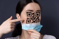 New reality. Close-up portrait of a woman taking off her mask and pointing at a face with a qr code. Dark background