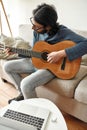 New skill. Young focused man sitting on sofa at home and playing acoustic guitar, watching online music course on laptop Royalty Free Stock Photo