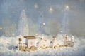 New Year card with toy train in a fairy village on winter background with snow and lights.