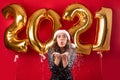 New Year celebration. Attractive woman in party dress and Santa hat blowing snow near balloons 2021, red background Royalty Free Stock Photo