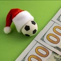 New Year`s soccer ball in a red Santa Claus hat next to the hundred dollar bills on a green background. Money, christmas and Royalty Free Stock Photo