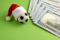 New Year sports betting. Souvenir soccer ball in a red Santa Claus hat next to dollars on a green background. The concept of a Royalty Free Stock Photo